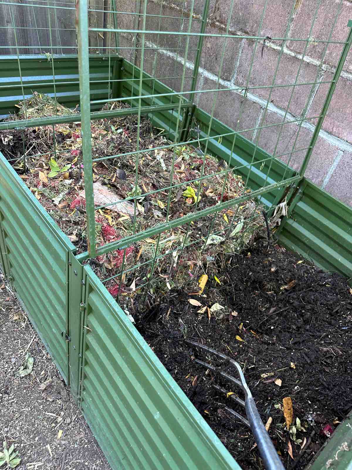 A green metal three bin compost system in the backyard with plant matter and compost at varying levels of decomposition.