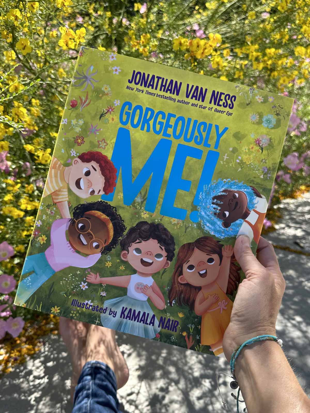 Holding out the children's book Gorgeously Me by Jonathan Van Ness in front of blooming spring flowers.