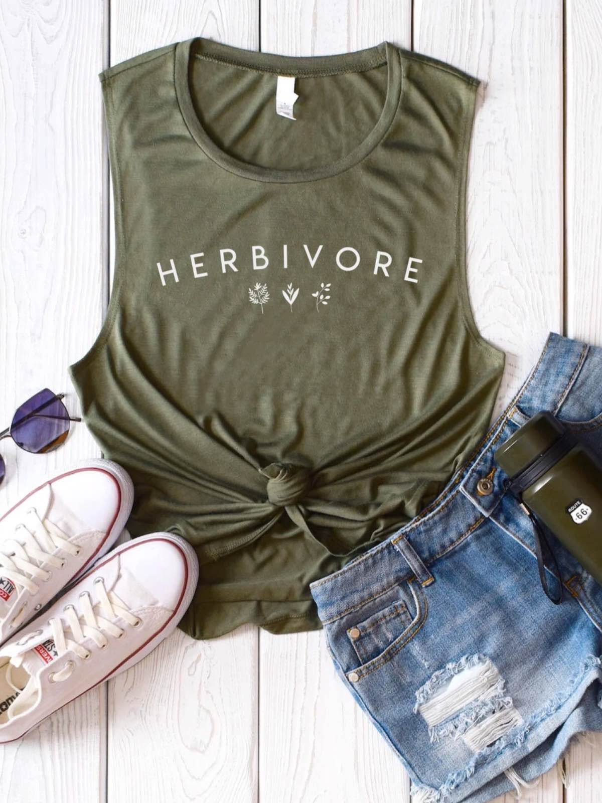 Outfit flatlay with a tank top that say "herbivore" next to vegan shoes, pants, and sunglasses.