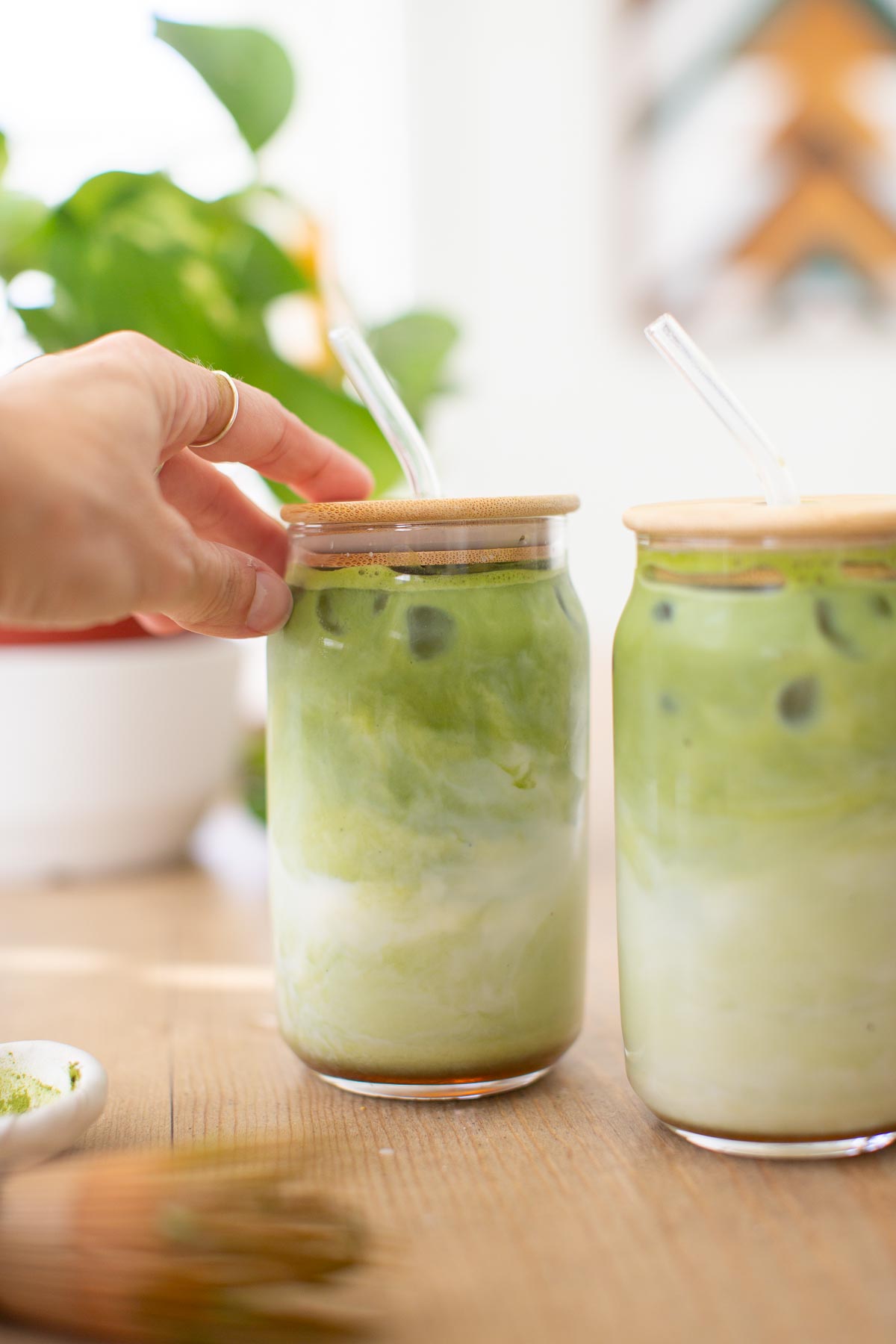 A hand reaching for an iced matcha latte glass with a wooden lid and glass straw.
