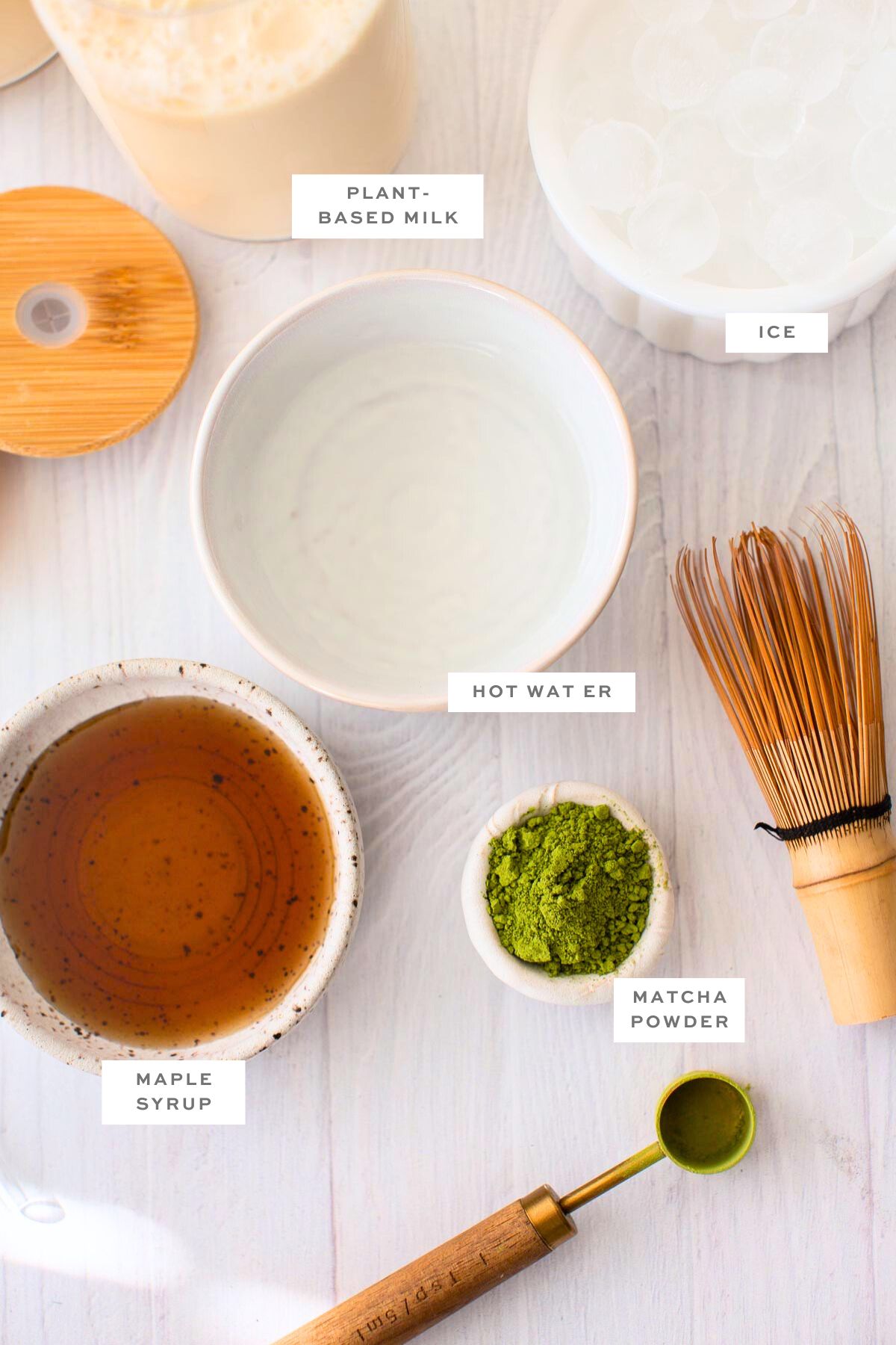 Gathered ingredients for making iced matcha lattes, including matcha powder, milk, ice, hot water, and maple syrup.