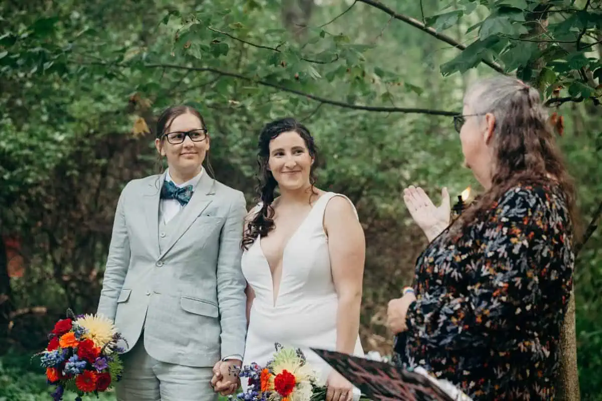 Queer brides exchanging vows in rainbow ceremony with mom officiating.