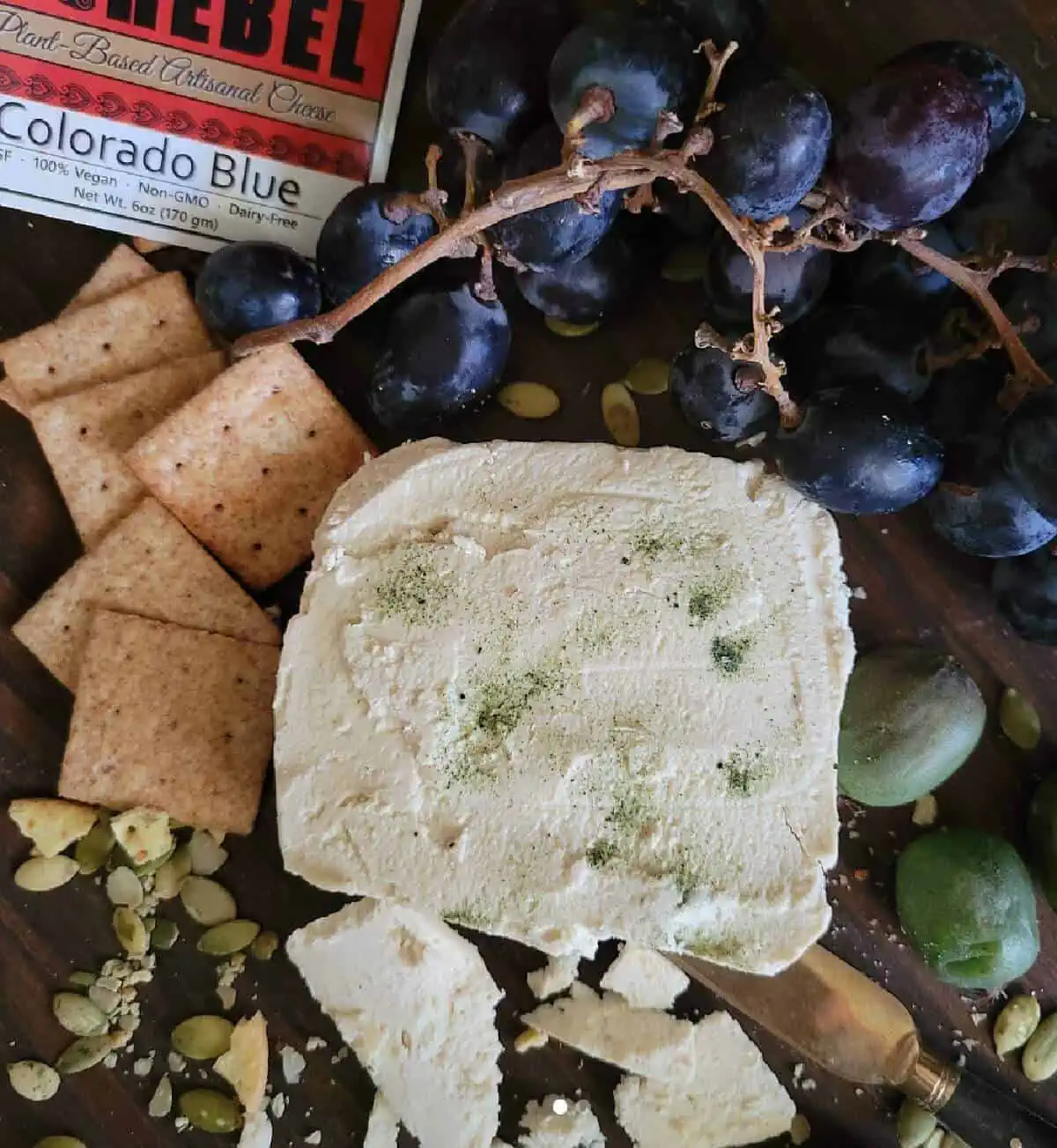 A large square of Peaceful Rebel vegan blue cheese laying on top of an assortment of crackers, grapes, pumpkin seeds, and olives along with the cheese wrapper and a cheese knife.