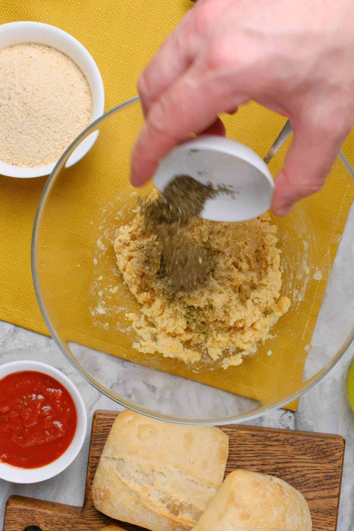 Seasonings being aded to a bowl of mashed chickpeas.