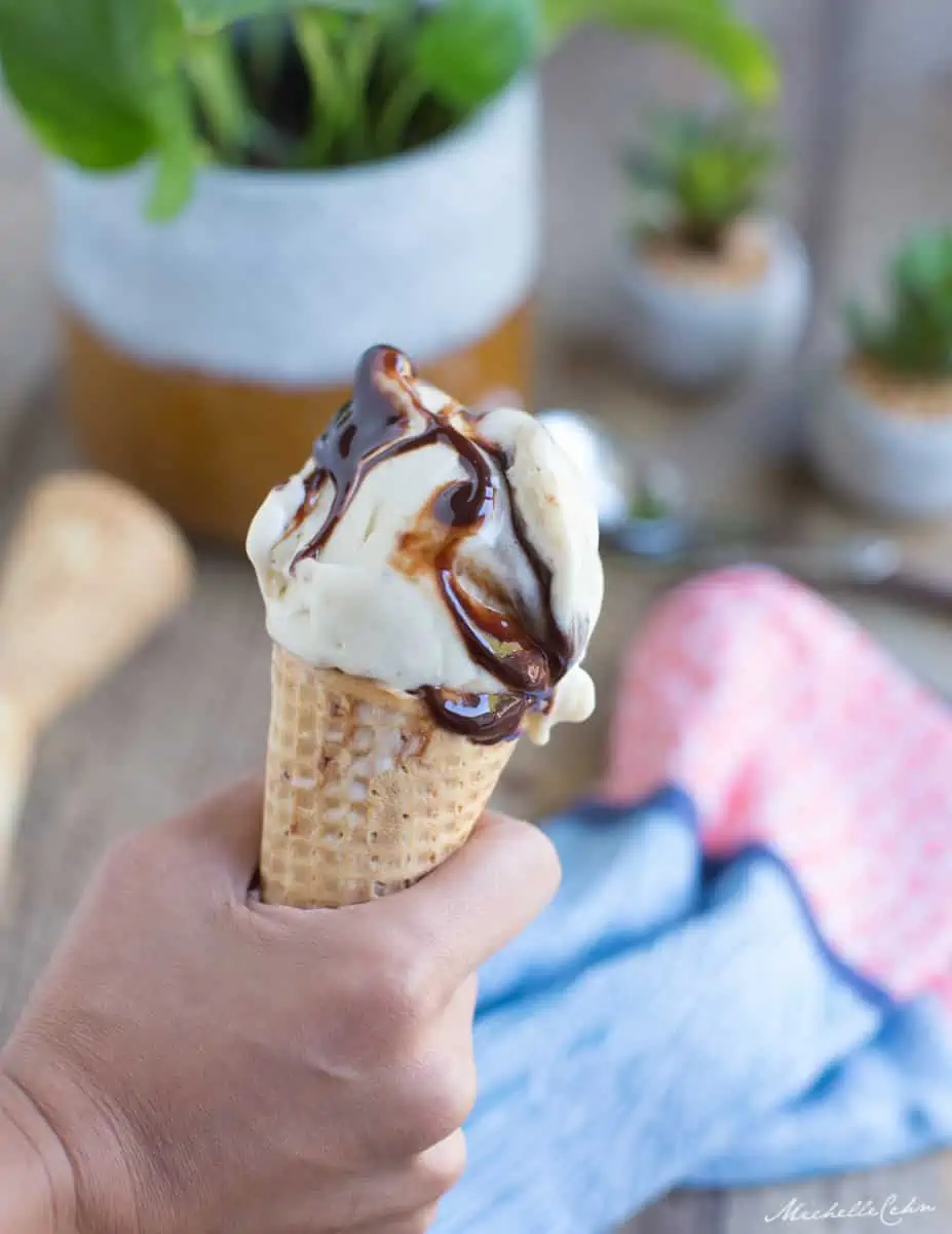 A hand holding a waffle cone topped with banana nice cream that has been drizzled with chocolate sauce.