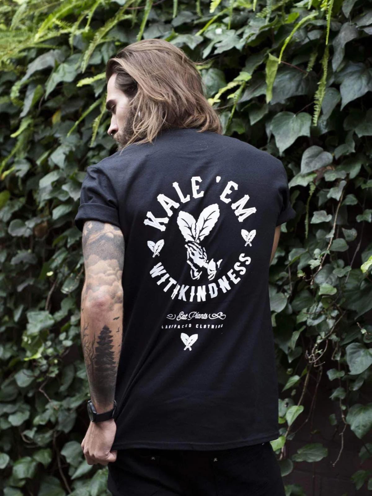 Tall vegan man outdoors wearing a "Kale 'em with kindness" black t-shirt from the brand Plant Faced Clothing.
