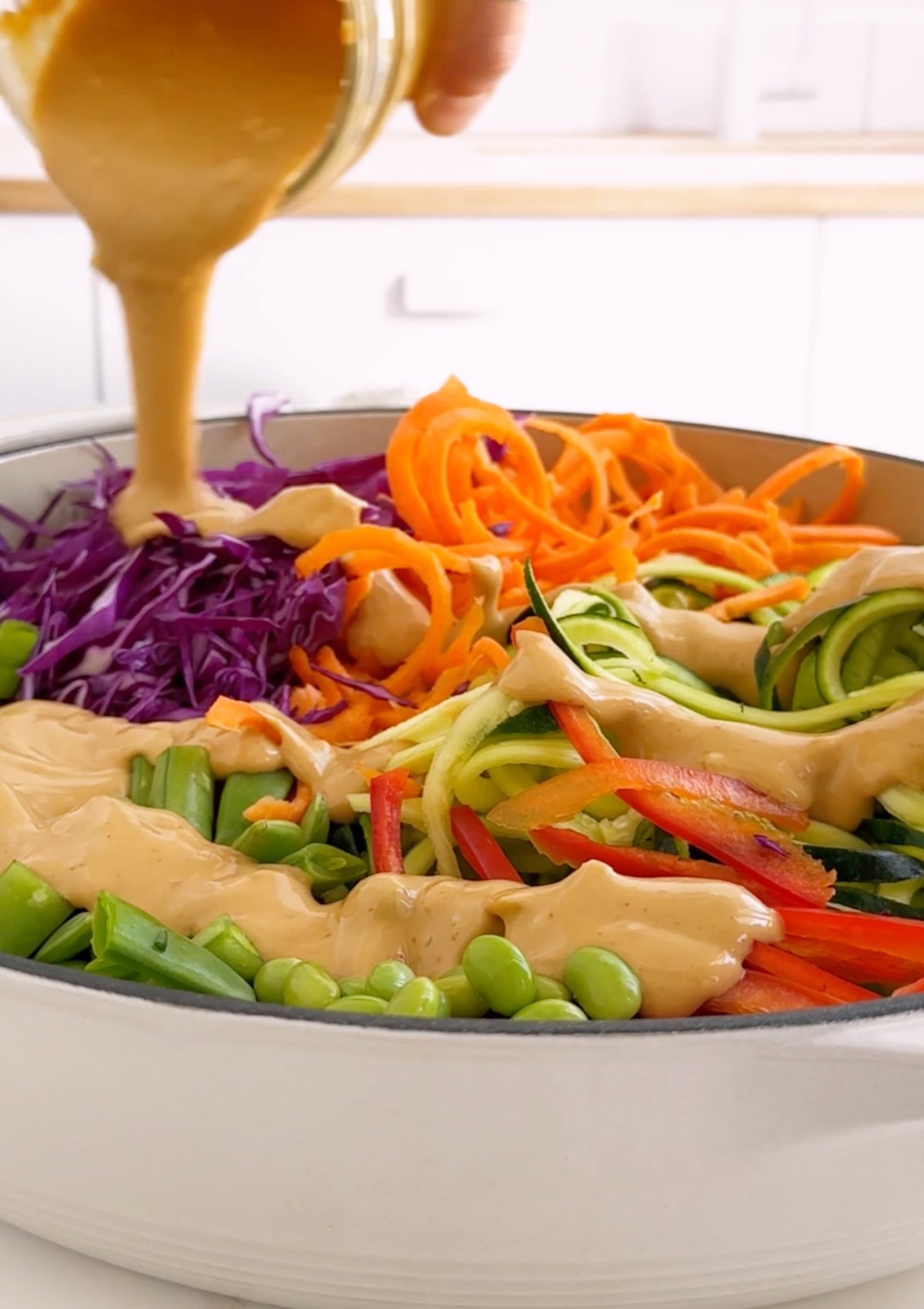 Pouring peanut dressing over bed of zucchini noodles, carrot noodles, and other vegetables.