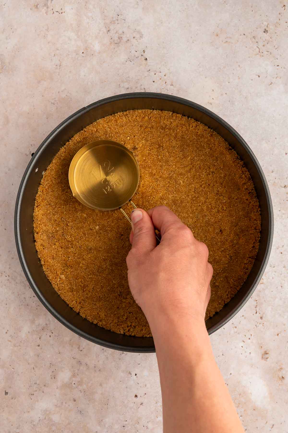 A hand pressing the vegan graham cracker crust into a springform pan with the back of a measuring cup to flatten it.