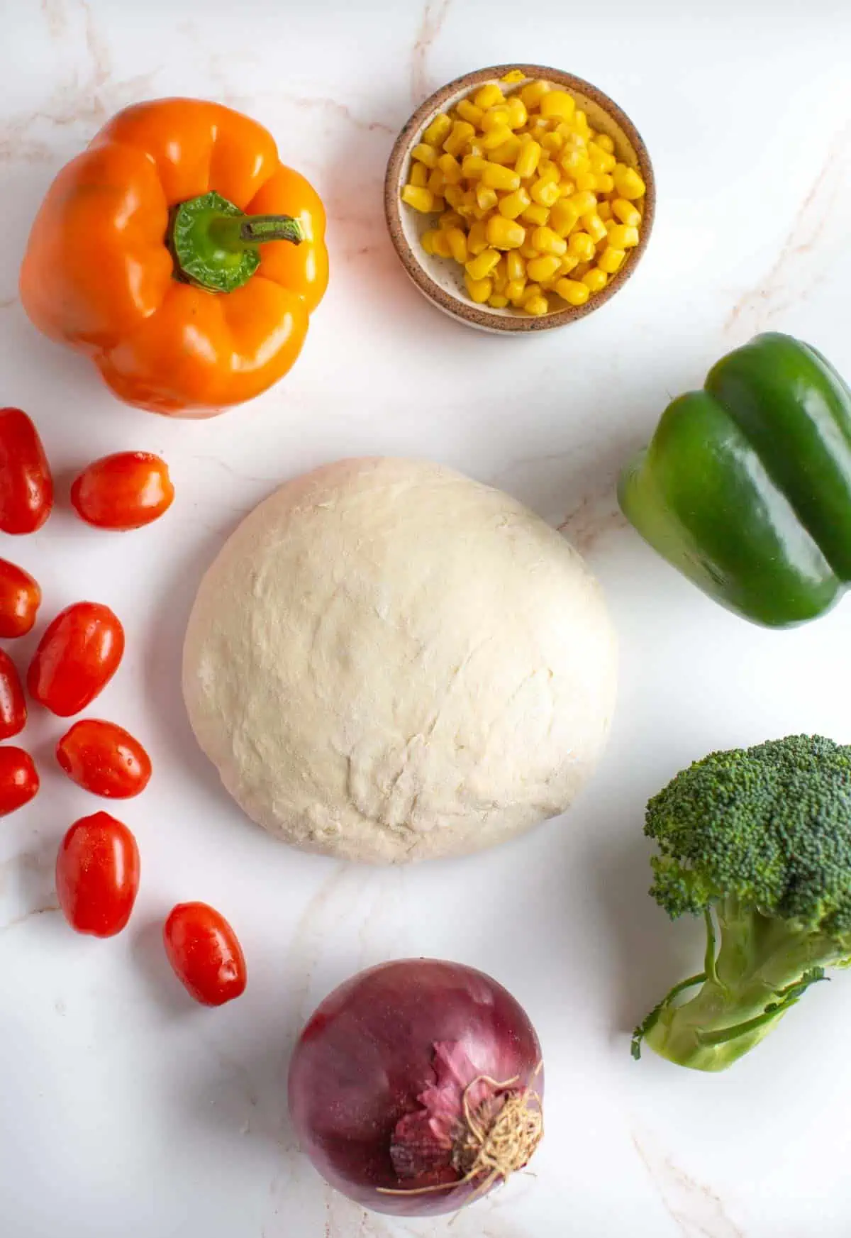 Photo of a ball of pizza dough surrounded by a rainbow of veggies including cherry tomatoes, bell pepper, corn, broccoli, and red onion.