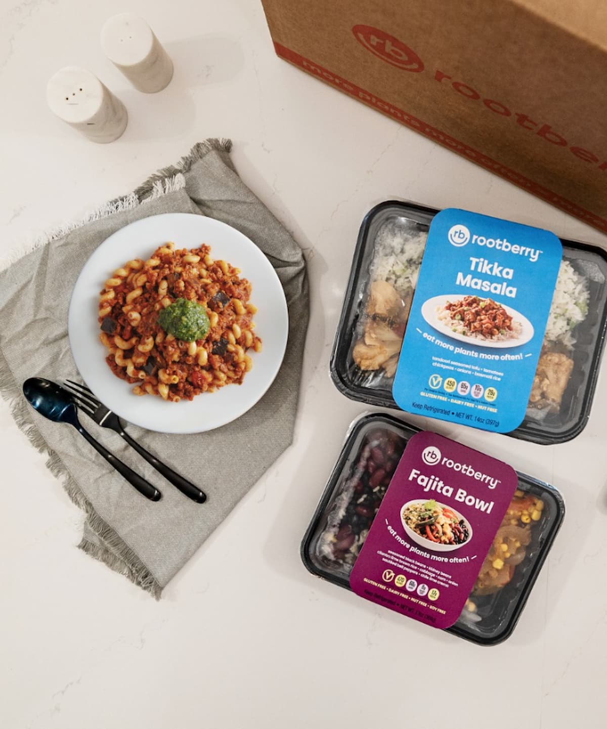 Flatlay of a prepared vegan pasta dish alongside Rootberry’s packaged Tikka Masala and Fajita Bowl meals, showcasing the brand’s ready-to-eat, plant-based meal delivery options.