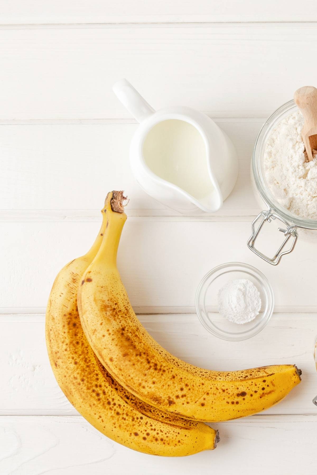 Bananas, flour, baking powder, all-purpose flour, and plant-based milk on a tabletop.