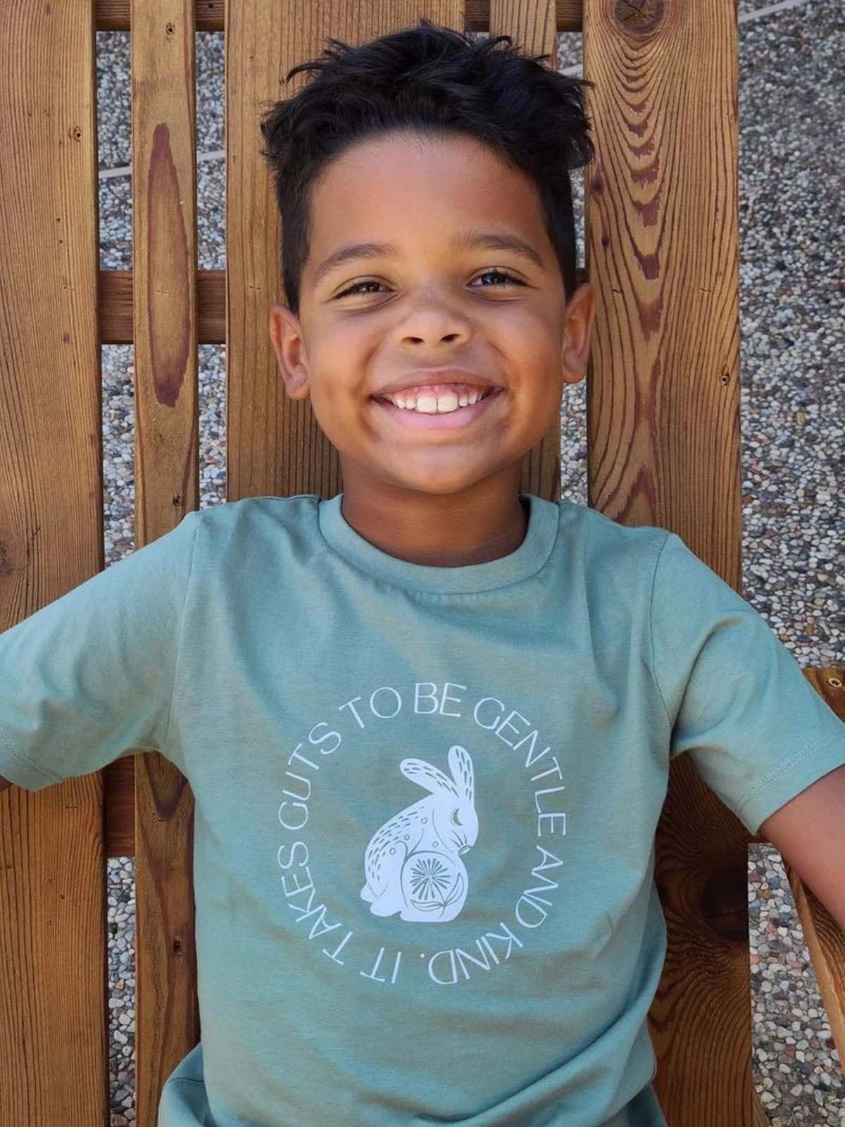 A vegan boy wearing a sage green shirt that says "it takes guts to be gentle and kind" from Small but Mighty Tees shop.