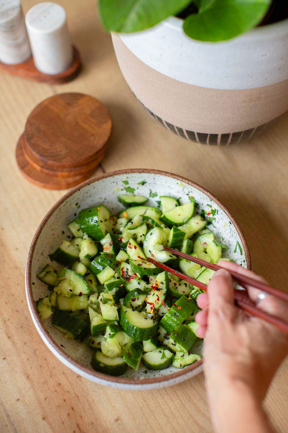 A hand reaching with chopsticks into a bowl of smashed cucumber salad with sesame and red pepper flakes.