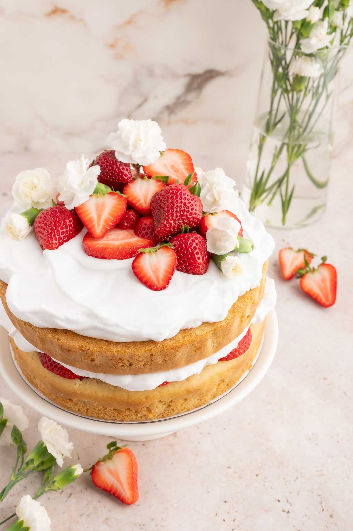 A round 2-layer vegan strawberry shortcake on a cake stand and topped with coconut whipped cream and strawberries.