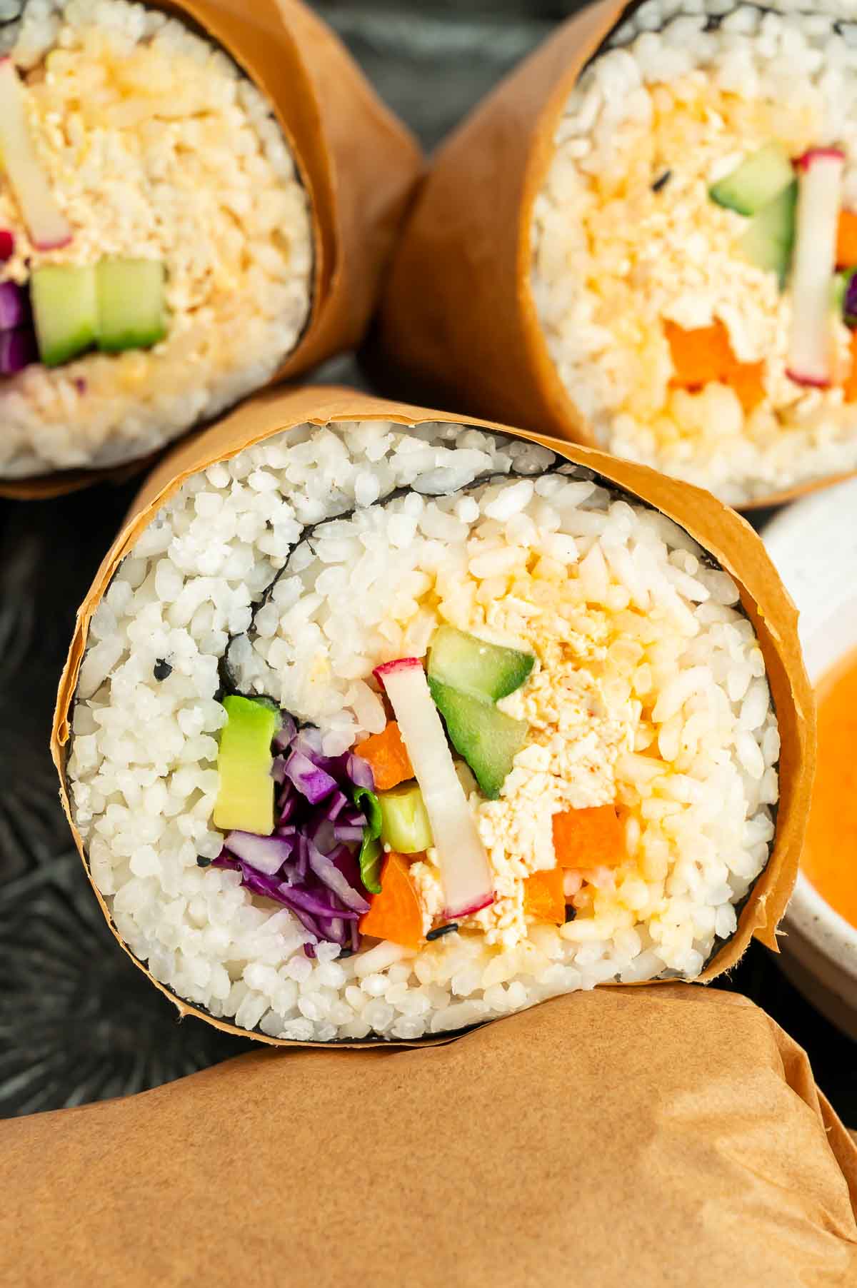 Close-up of colorful tofu sushi burritos neatly arranged on a dark tray with dipping sauce, wrapped in parchment and filled with fresh vegetables and seasoned rice.