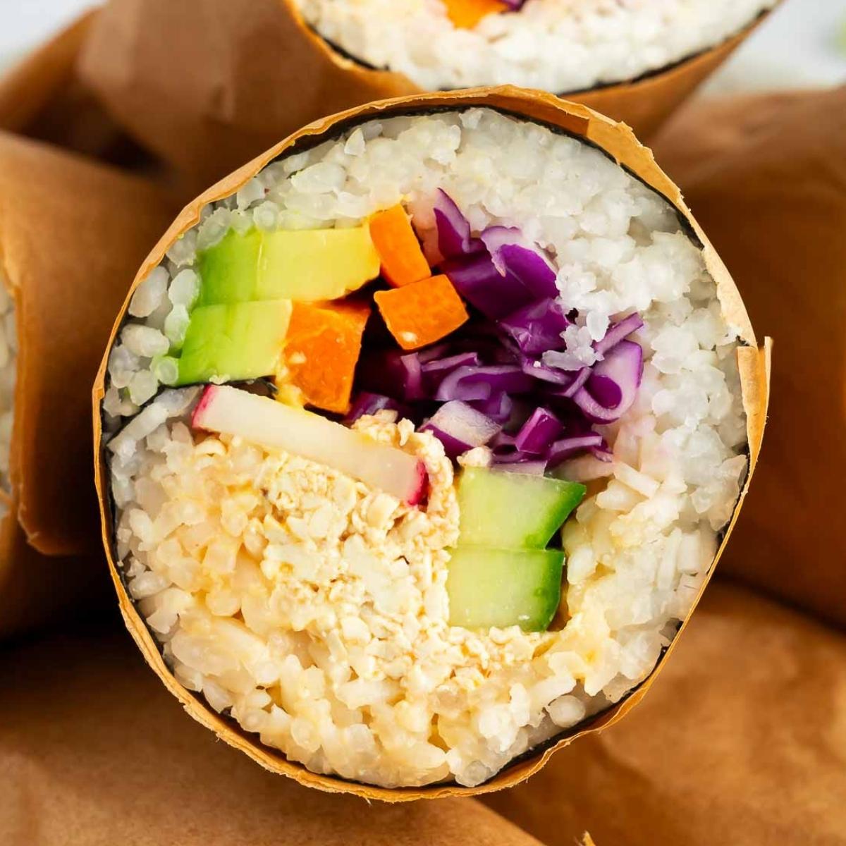 A vibrant vegan sushi burrito wrapped in brown paper, showing layers of sushi rice, avocado, shredded tofu, radish, purple cabbage, cucumber, and carrot.