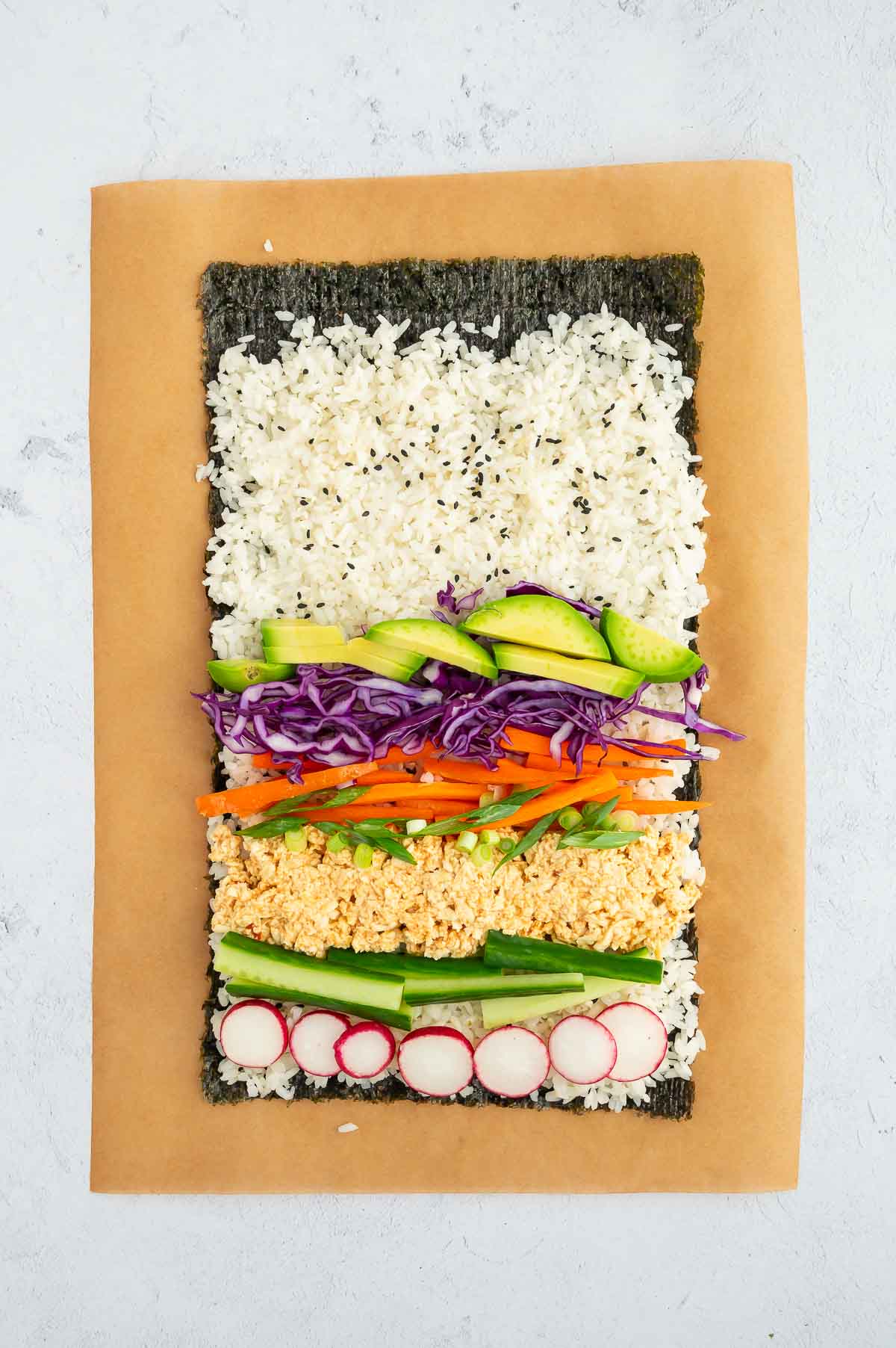 Nori sheet topped with seasoned sushi rice, avocado, shredded tofu, purple cabbage, radish, and carrots, ready to be rolled into a sushi burrito.