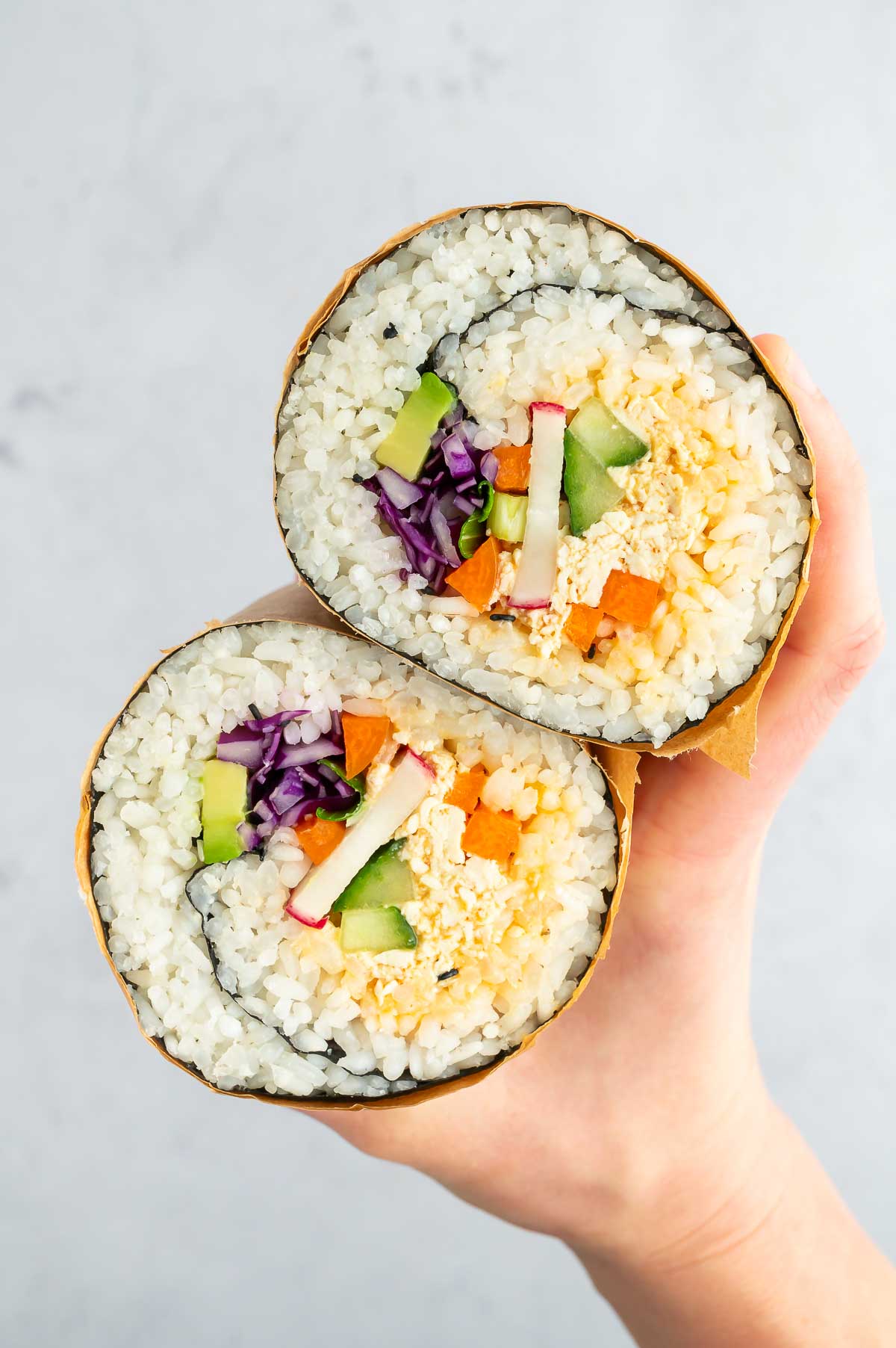A hand holding a sliced vegan sushi burrito, revealing vibrant layers of tofu, cucumber, cabbage, radish, and sushi rice.