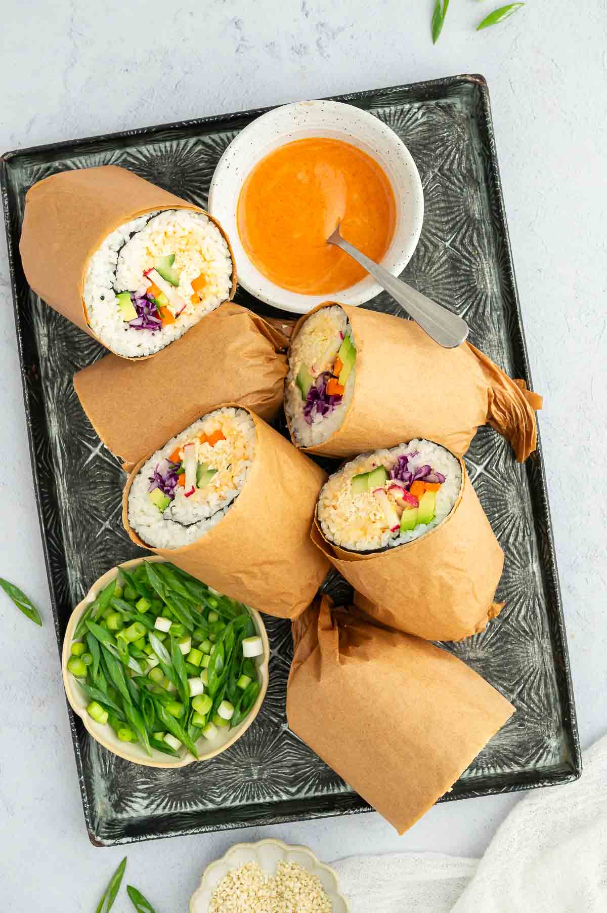 Sushi burrito halves wrapped in parchment arranged on a platter with spicy dipping sauce and a bowl of chopped scallions on the side.