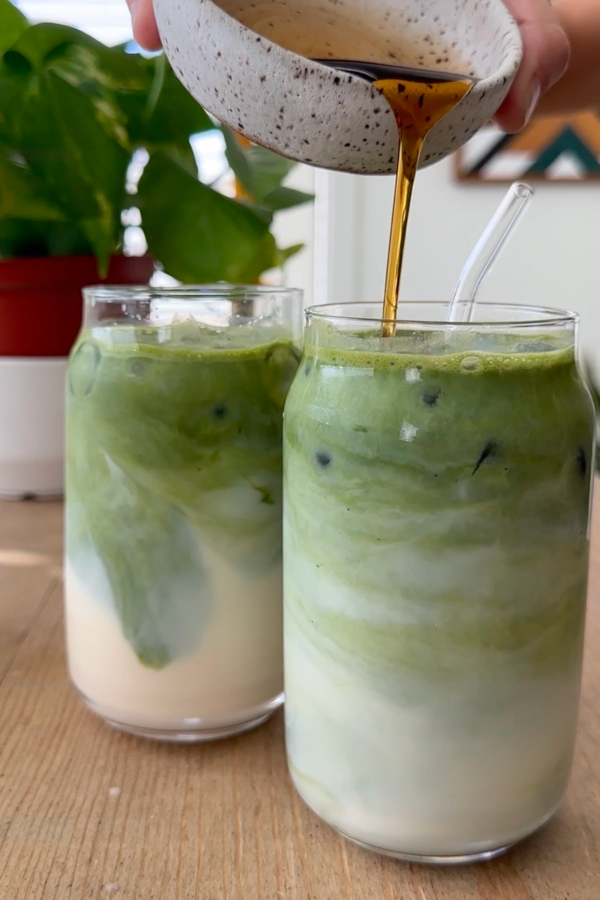 Maple syrup pouring from a small bowl into the glasses of prepared iced matcha lattes.