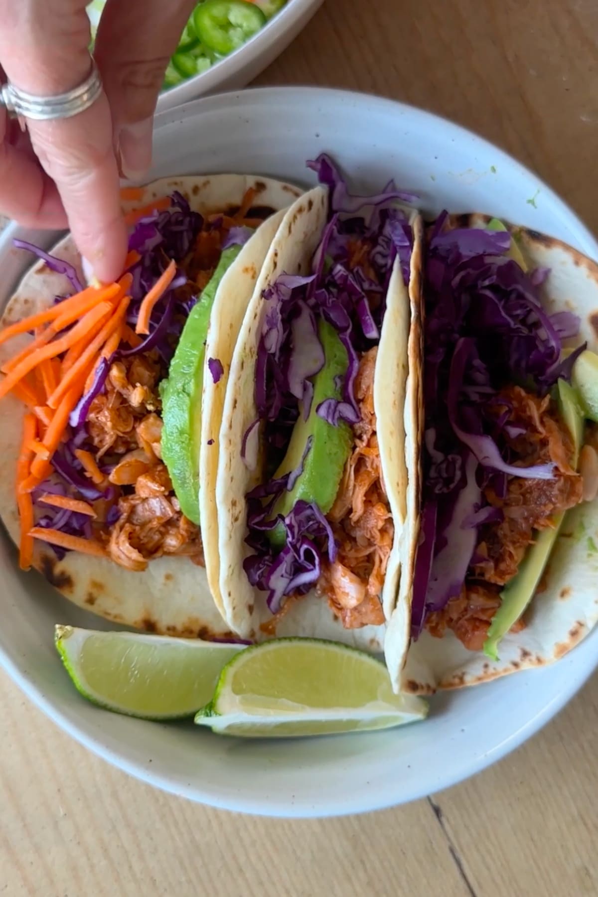 A hand sprinkling shredded carrots on top of bbq jackfruit tacos.