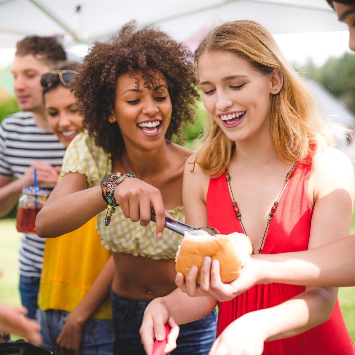 Friends laughing and serving food at an outdoor vegan BBQ, holding a sandwich bun and tongs under a sunny canopy.