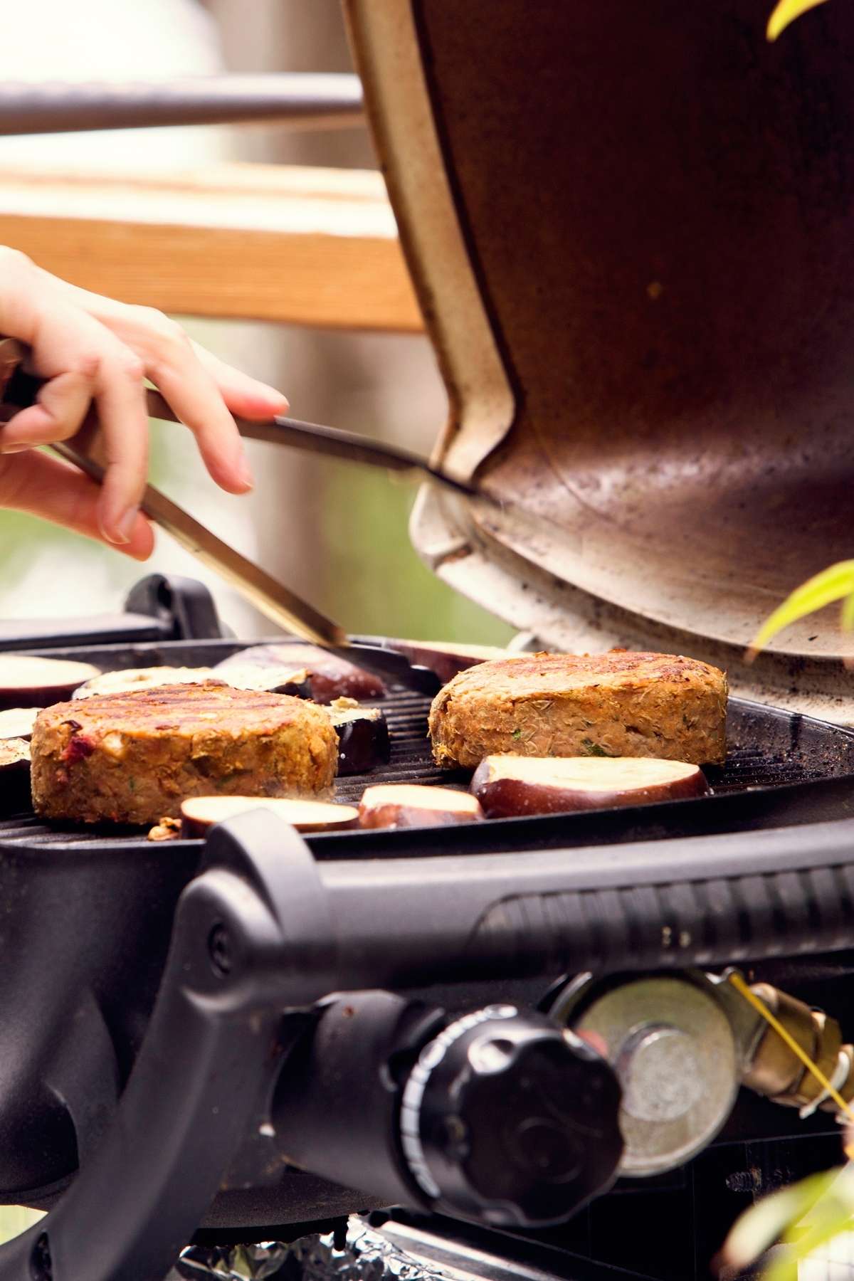 Close-up of vegan burger patties and sliced eggplant grilling on a gas barbecue, with tongs flipping the food under an open lid.