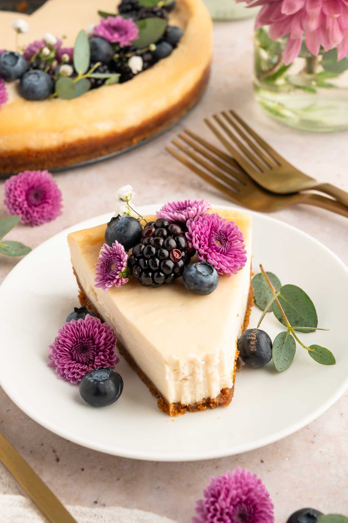 A slice of vegan cheesecake on a plate with a bite missing and topped with blueberries, blackberries, and edible flowers.