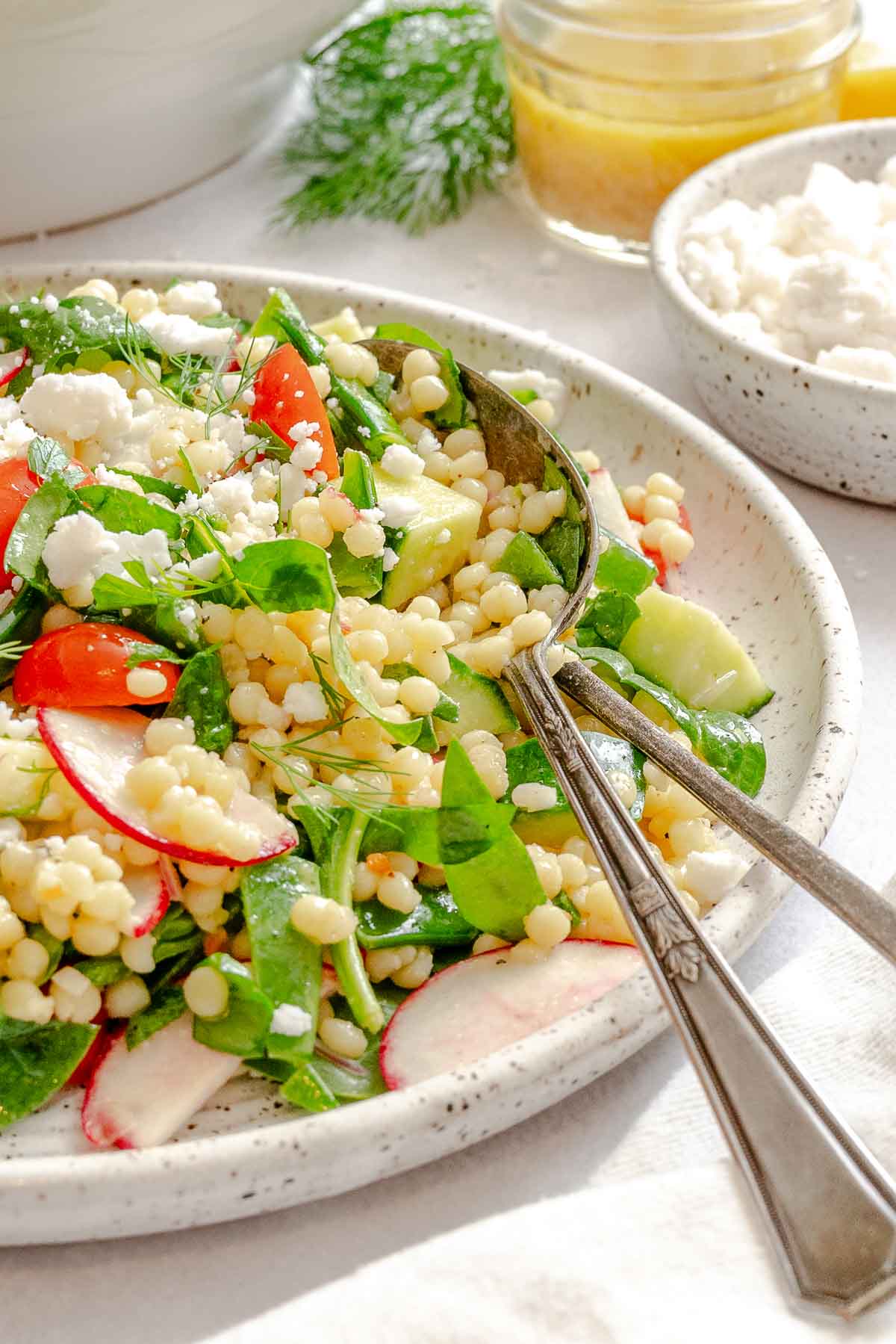 Vibrant vegan couscous salad with cherry tomatoes, cucumber, and spinach in a speckled bowl with vinaigrette on the side.