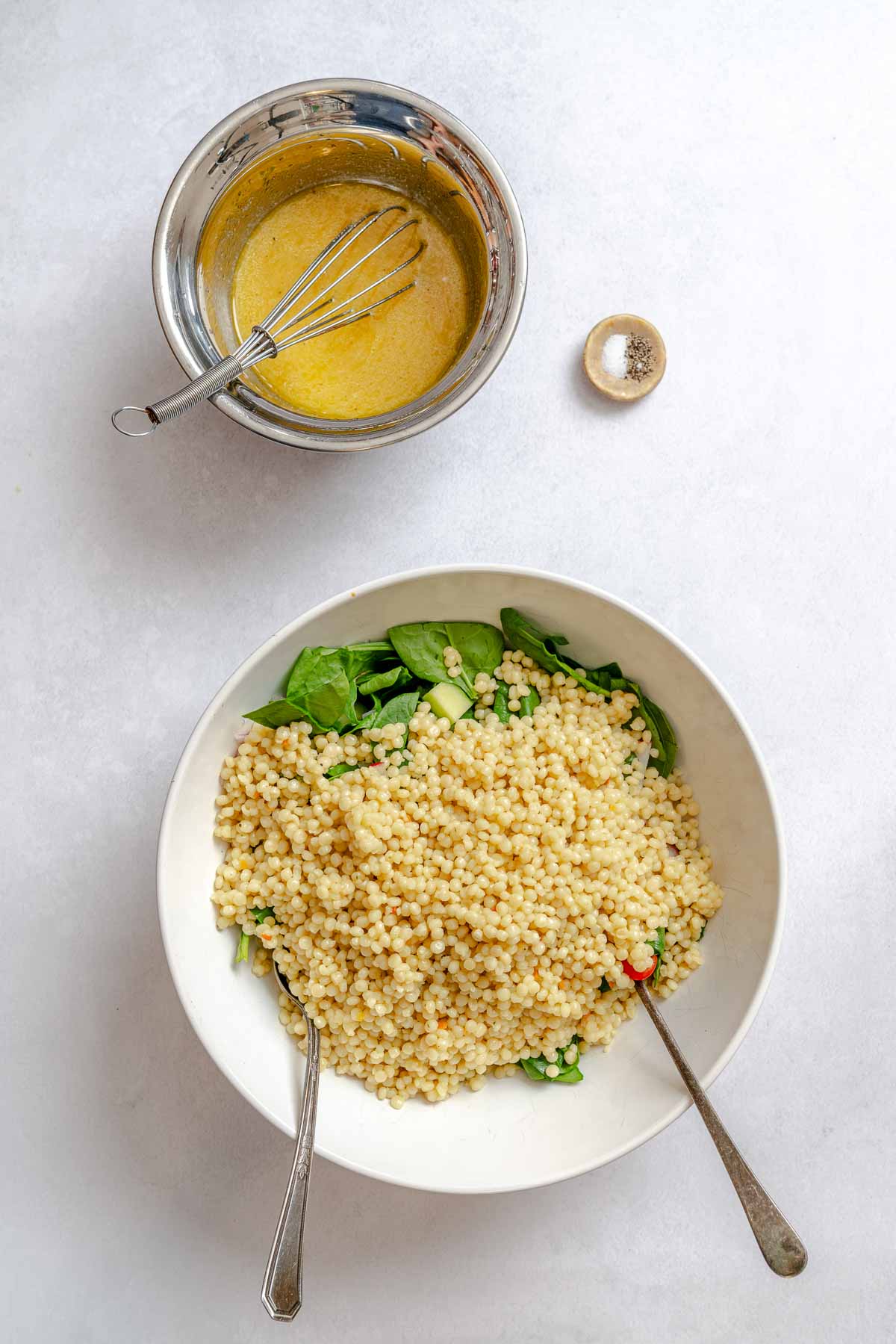 Cooked Israeli couscous and chopped spinach in a mixing bowl next to a whisked lemon vinaigrette dressing.