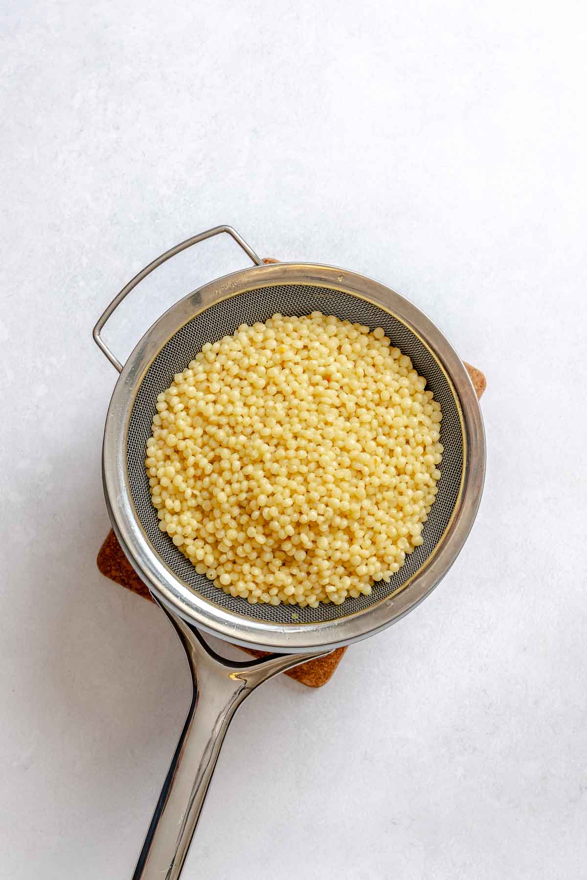 Cooked pearl couscous draining in a fine mesh strainer after boiling, prepped for vegan couscous salad.