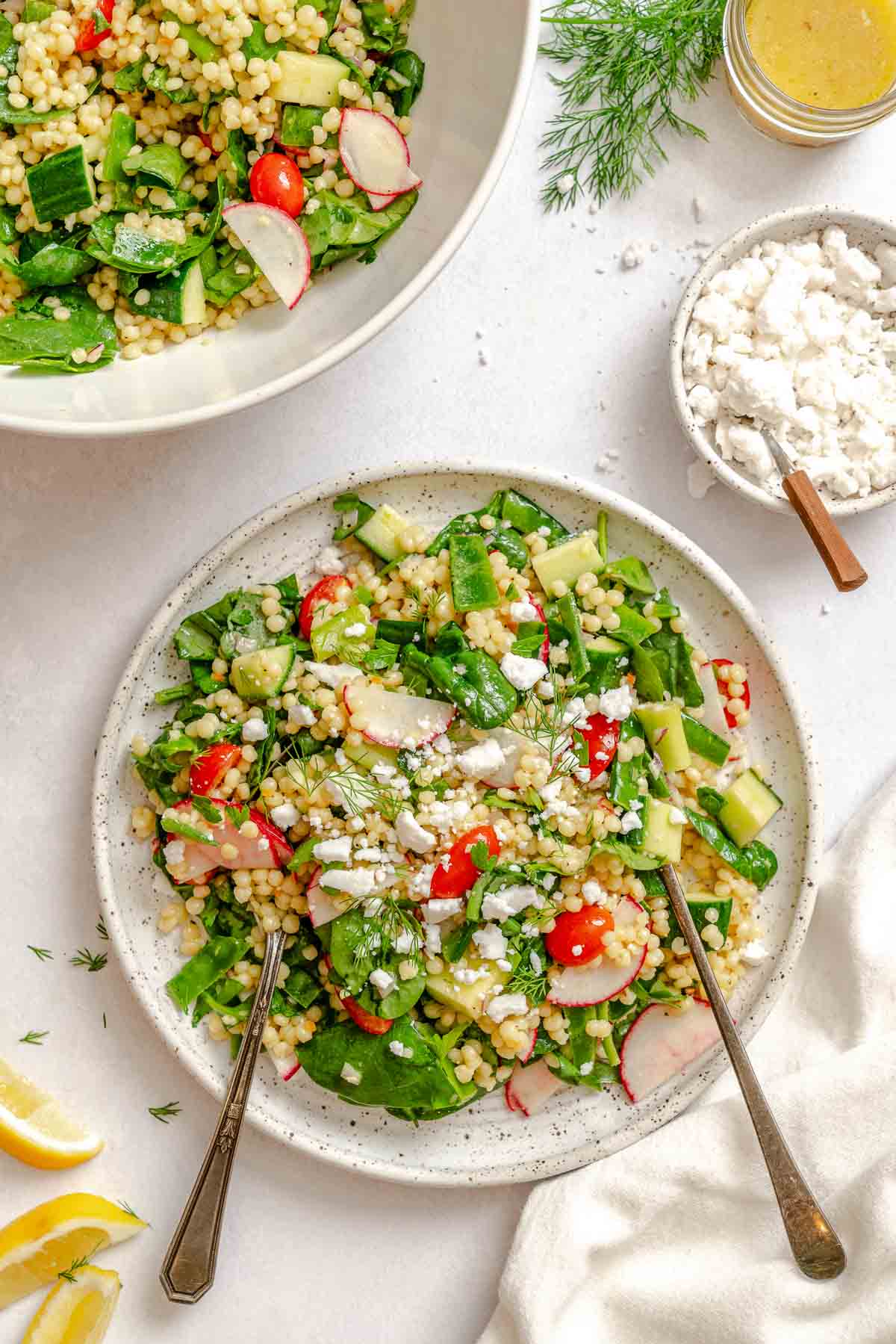 Two servings of fresh vegan couscous salad with spinach, cucumbers, and herbs on ceramic plates, ready to eat.