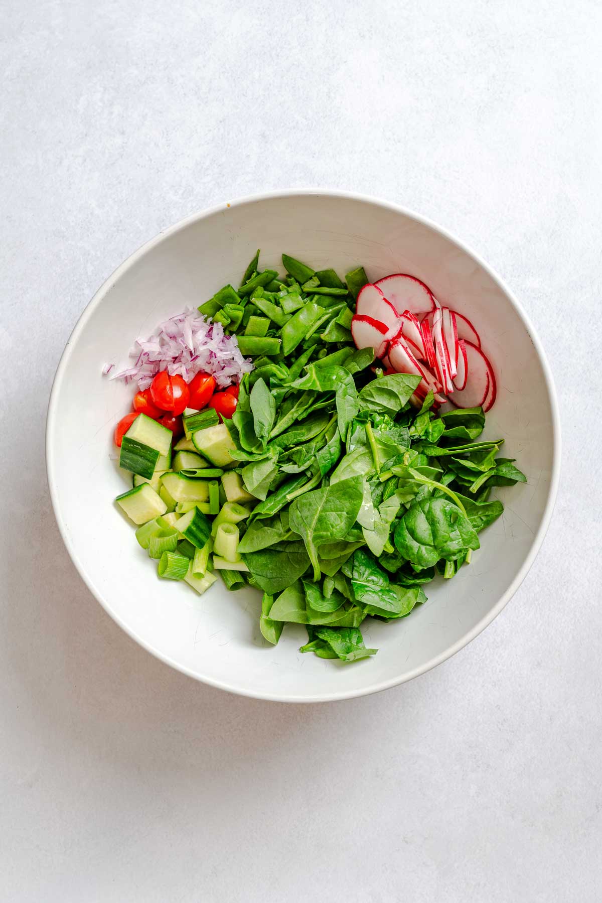 Chopped spinach, radish, cucumber, and red onion arranged in a large white bowl for couscous salad prep.