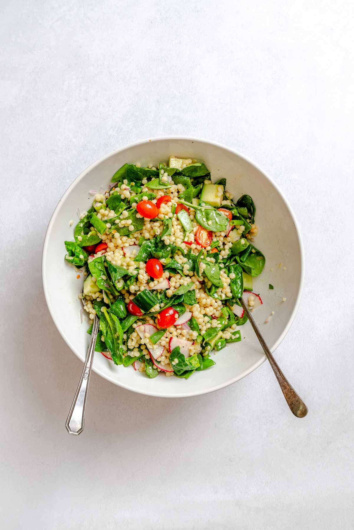 Tossed vegan couscous salad in a white bowl with spinach, cherry tomatoes, radish, and cucumber, ready to serve.