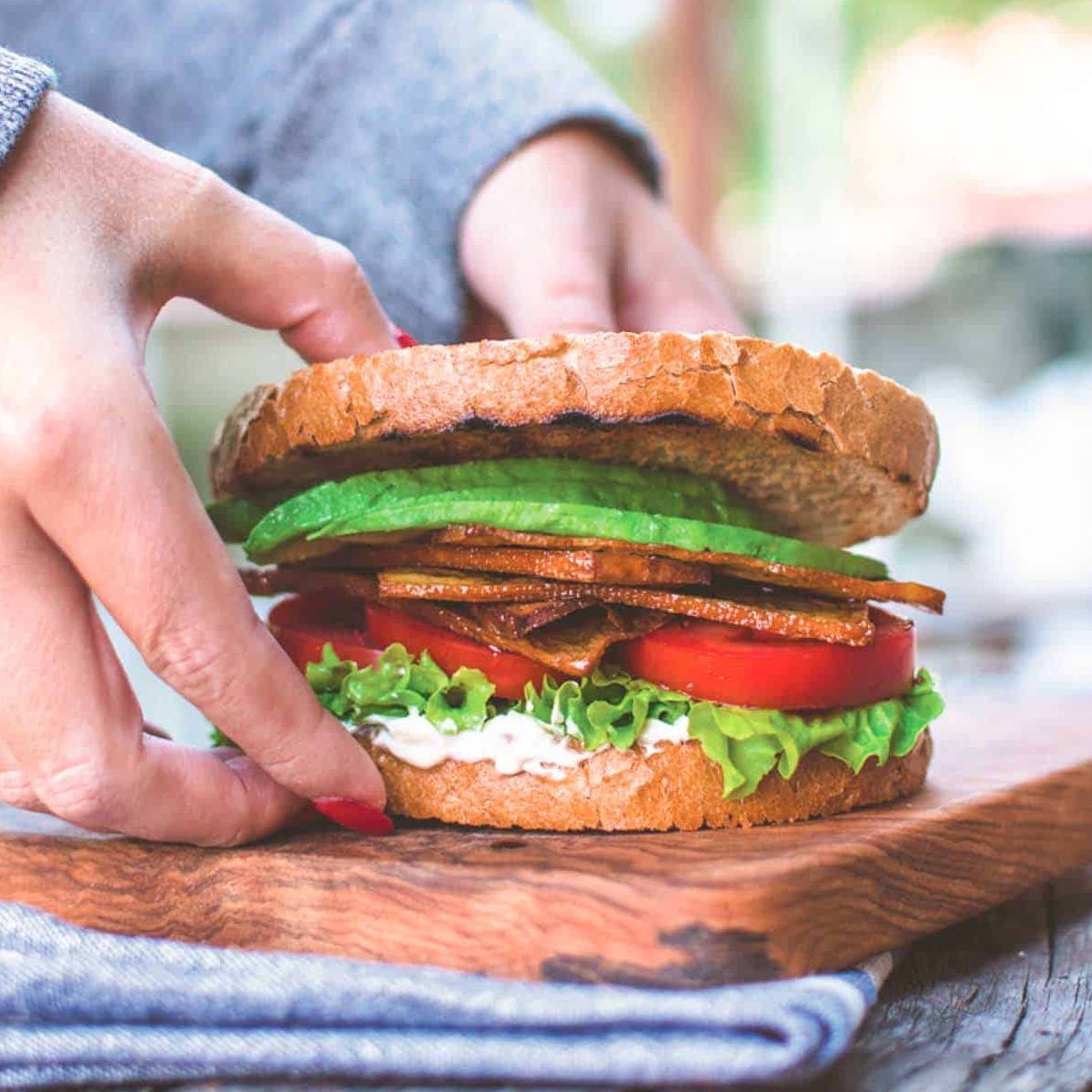 A woman holding a sandwich with crusty bread, lettuce, tomato, avocado, and vegan deli meat.