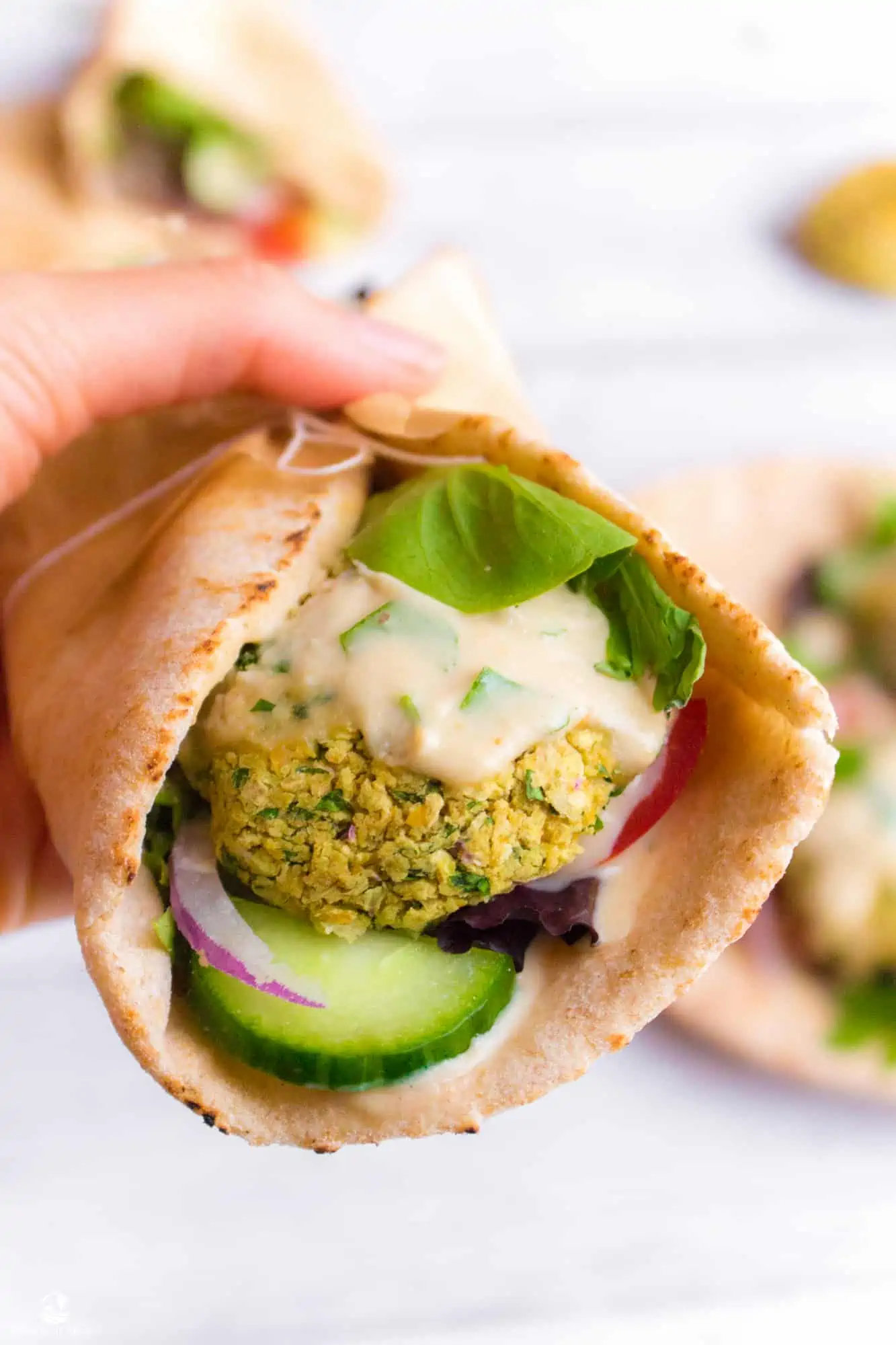 A hand holding a falafel pita sandwich stuffed with baked falafels, fresh vegetables, and a tahini lemon parsley dressing.