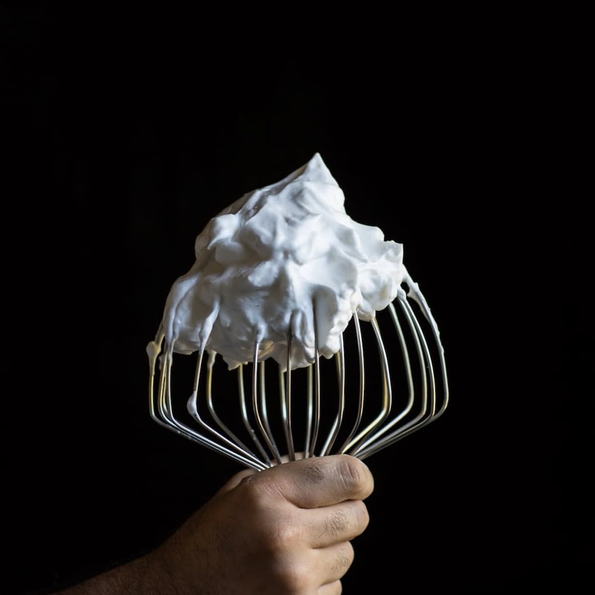 A hand holding a large whisk attachment covered in stiff, glossy vegan meringue made from aquafaba, set against a black background.