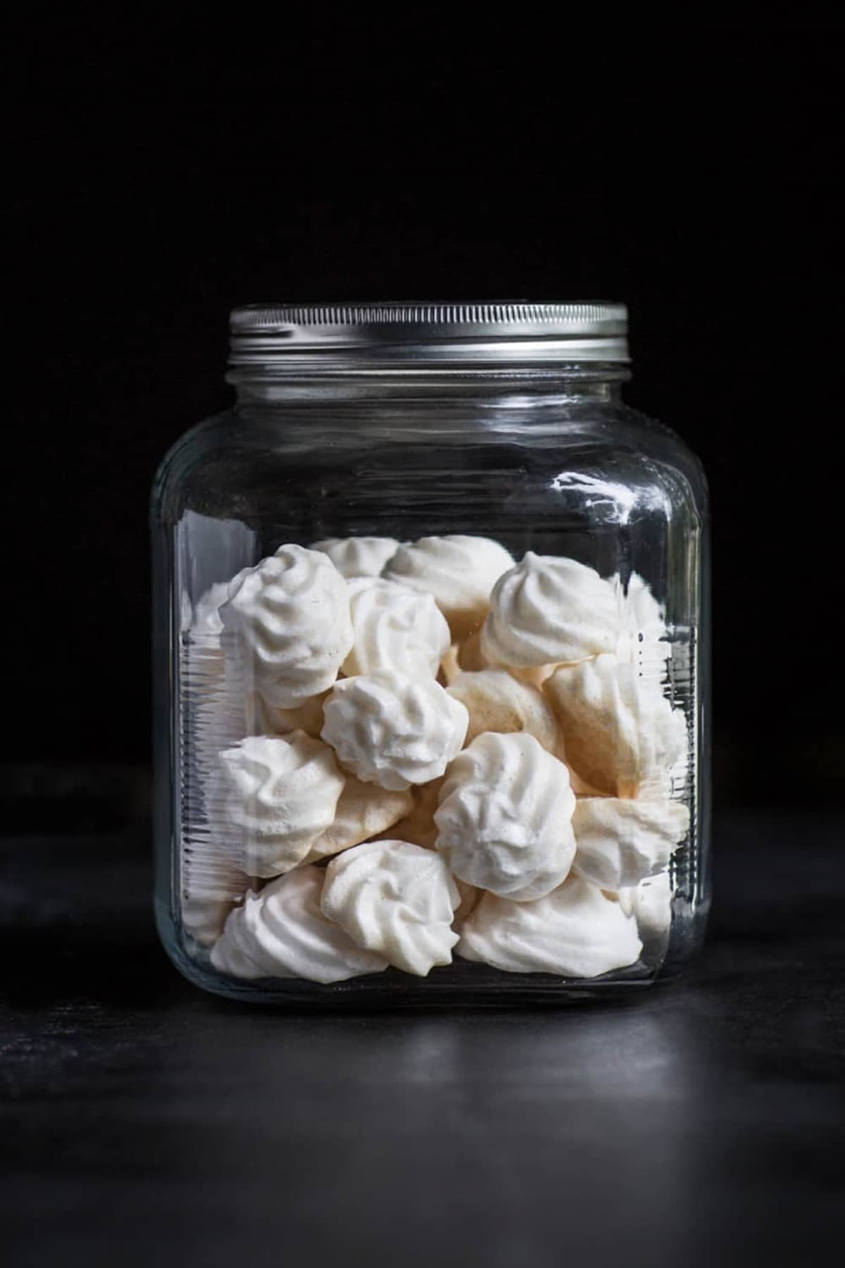 Crisp white vegan meringue cookies stacked in a clear glass jar with a silver lid, set against a dark background.