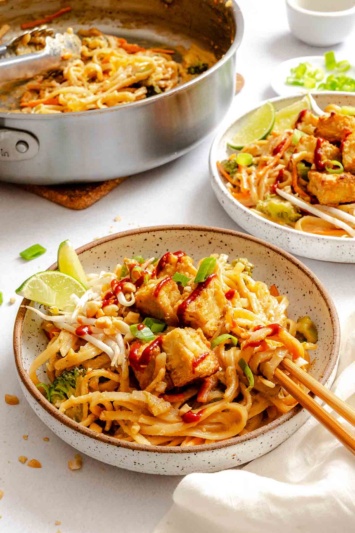 Vegan pad Thai served in bowls with tofu and lime, next to a skillet of noodles.