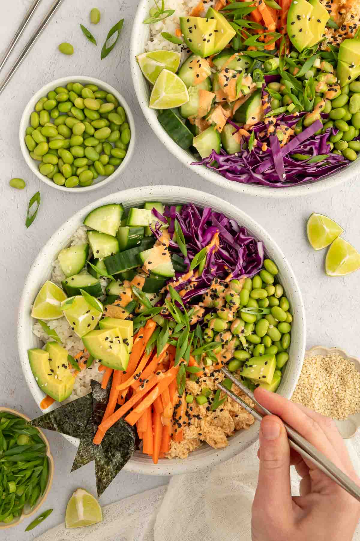 Hand holding chopsticks over a vegan poke bowl filled with rice, tofu, avocado, edamame, cabbage, and fresh vegetables.