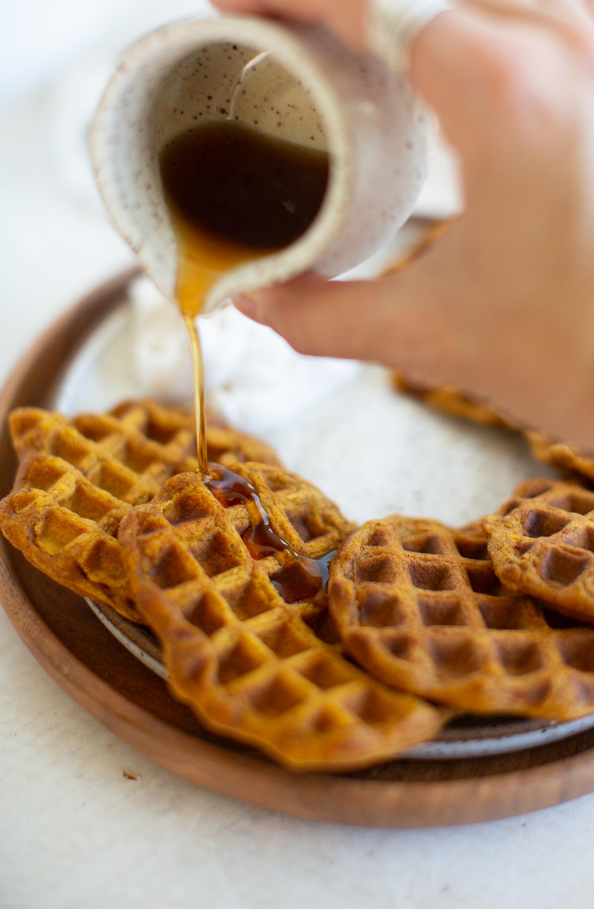 A hand pouring maple syrup from a small pitcher over a plate of vegan pumpkin waffles.