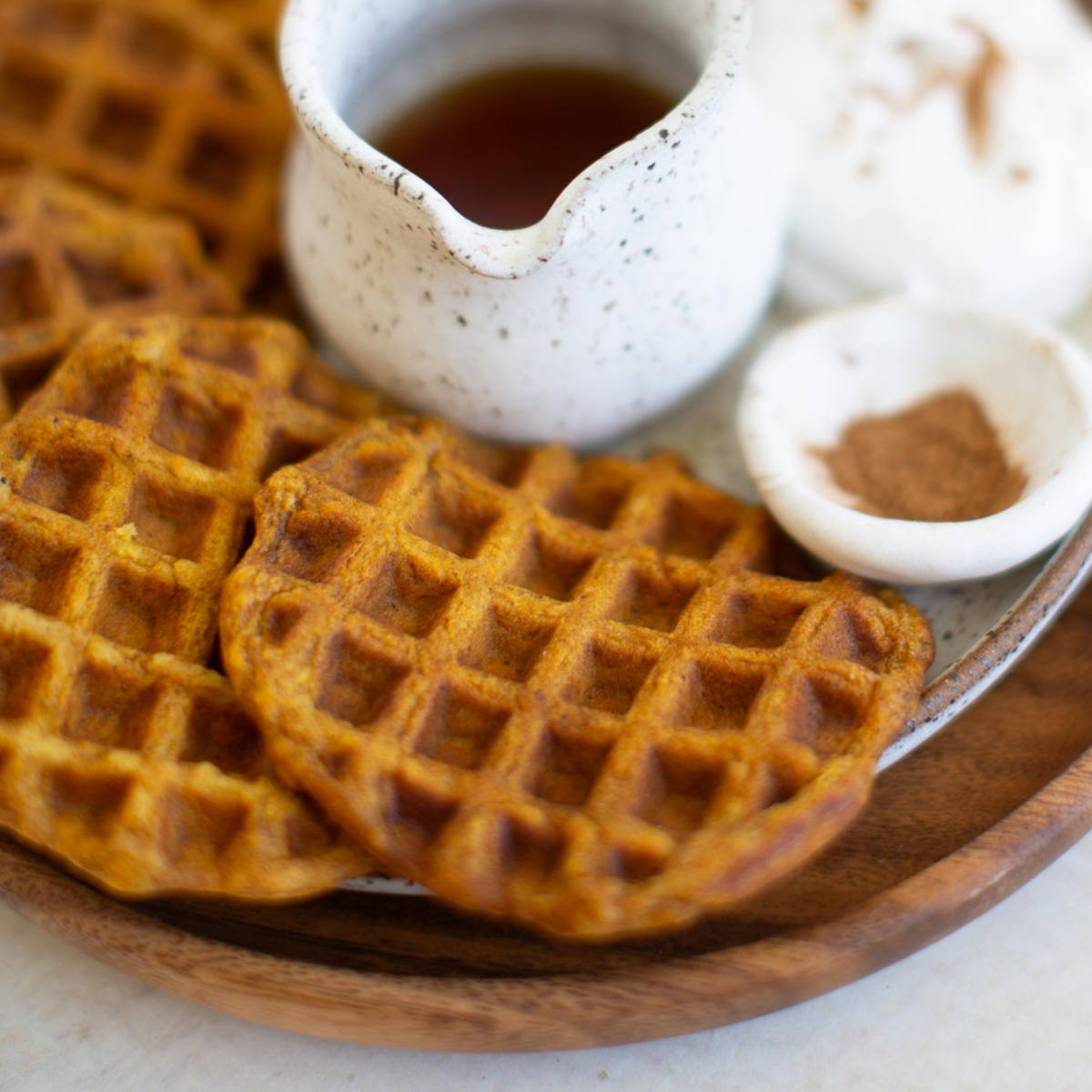 Vegan pumpkin waffles arranged on a platter with small pitcher of maple syrup and a small bowl of pumpkin spice.