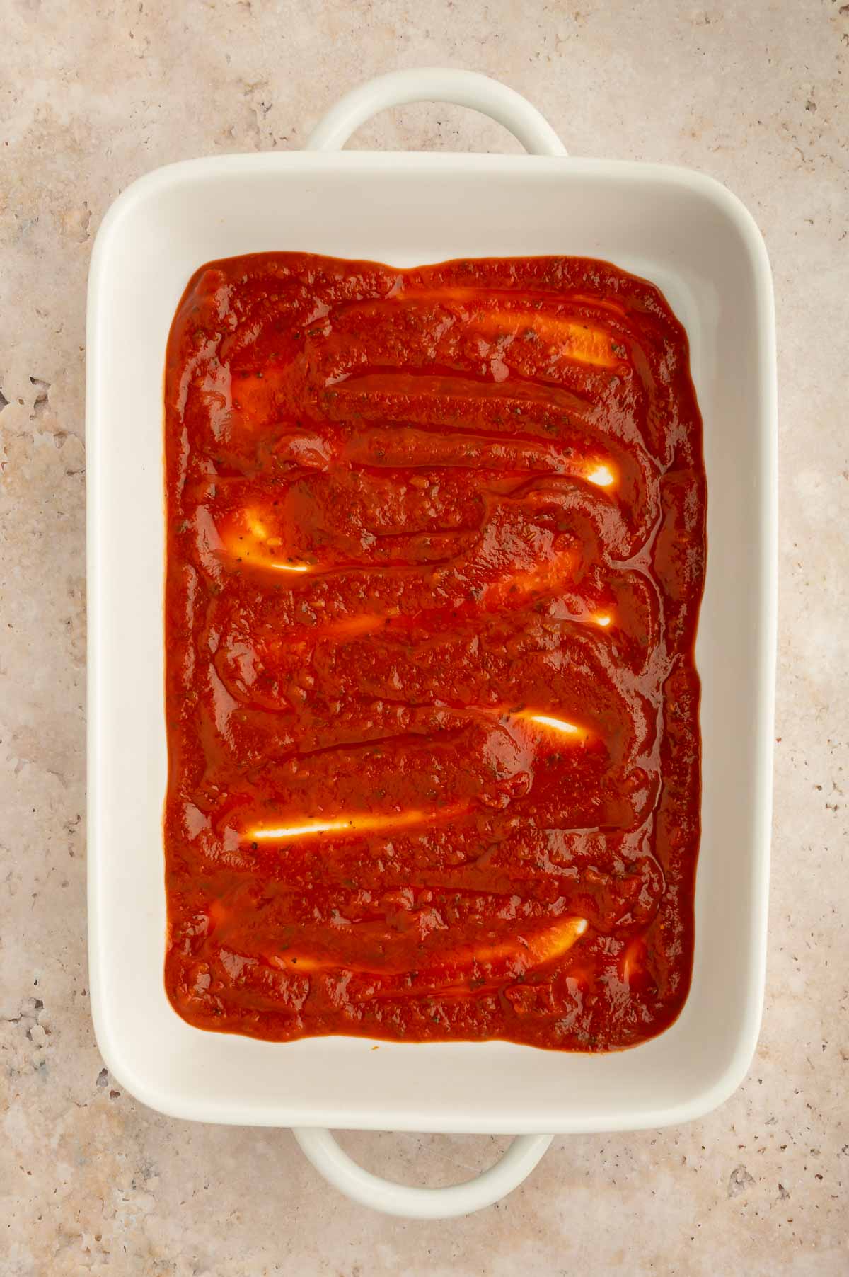 A casserole dish with marinara sauce spread on the bottom.