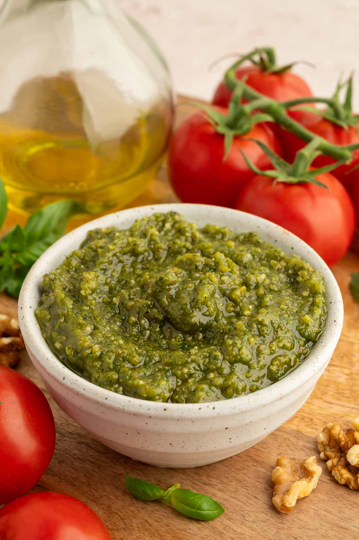 Vegan walnut pesto in a small white bowl, surrounded by walnuts, basil, and tomatoes.