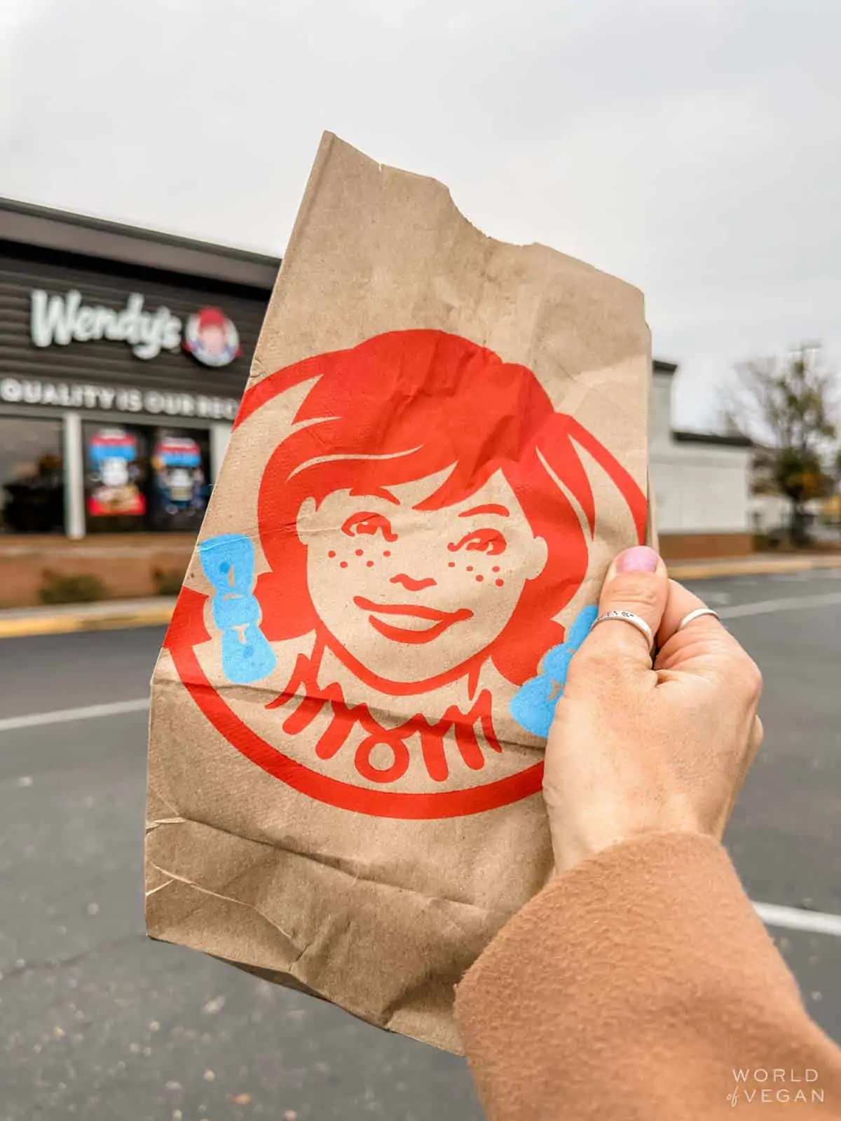 A hand holding up a Wendys paper bag in front of the Wendys restaurant.