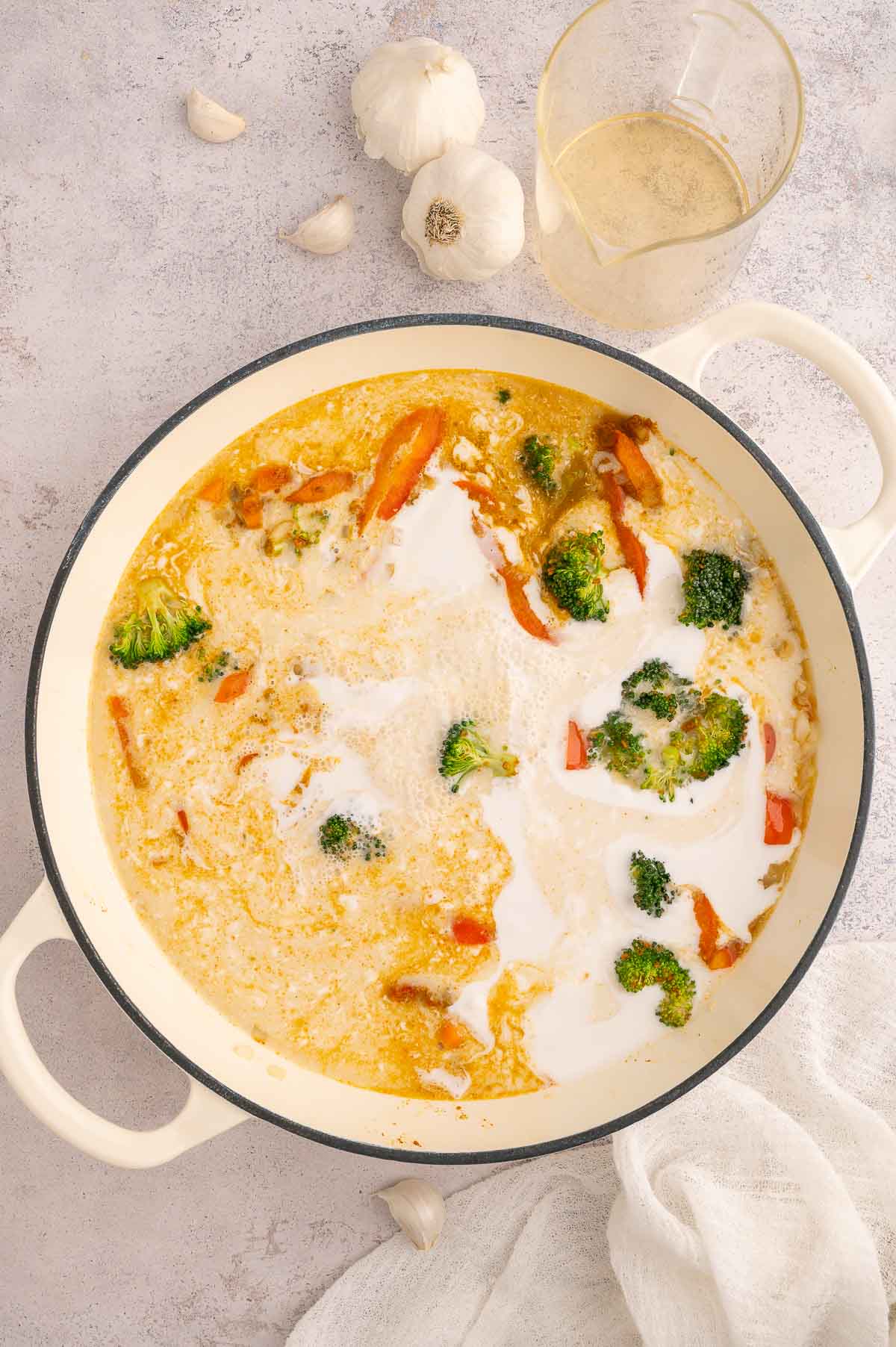 Coconut milk added to Thai curry soup with vegetables in a cream-colored pot.