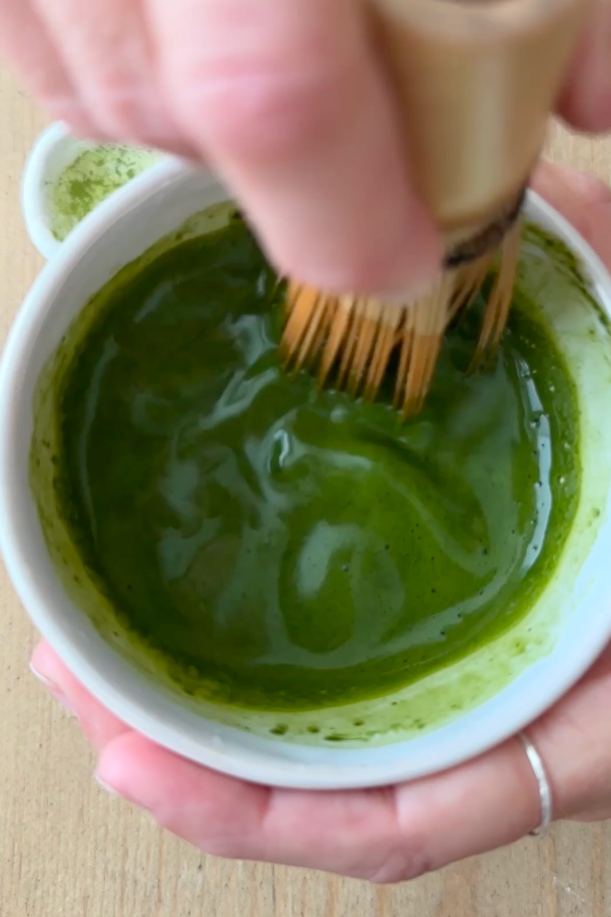 A hand using a bamboo whisk to mix together matcha powder and hot water in a small bowl.