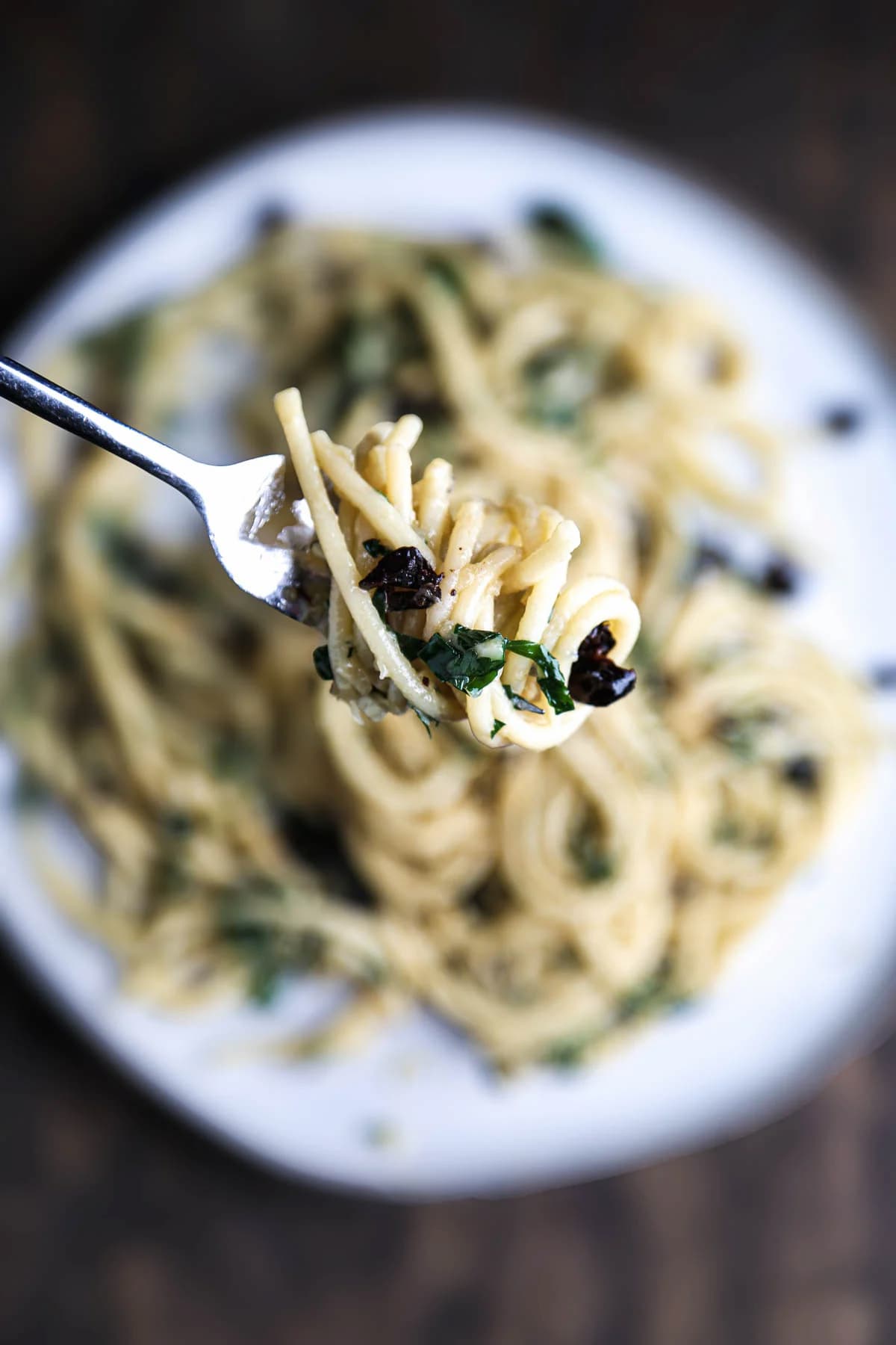 Fork twirling bucatini pasta coated in vegan white wine garlic sauce with parsley and olives.
